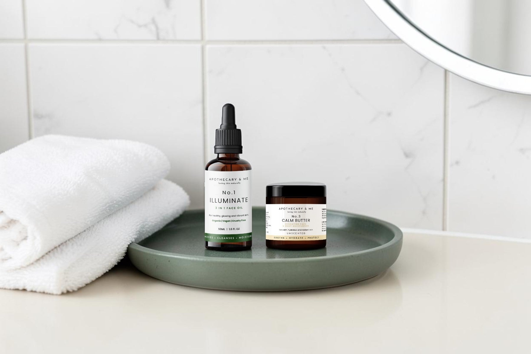Apothecary & Me No.1 Illuminate Face Oil and No.3 Calm Butter displayed on a sage green ceramic tray on a bathroom countertop, natural skincare products for dry and flaky skin.