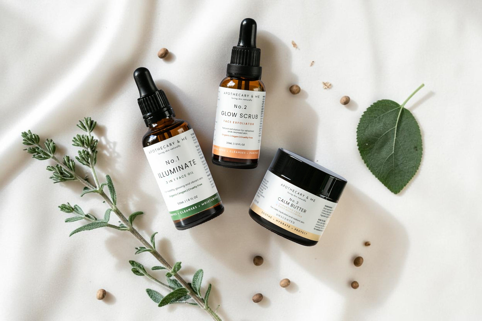 Apothecary & Me No.1 Illuminate Face Oil, No.2 Glow Face Scrub, and No.3 Calm Butter arranged on soft cream fabric with fresh herbs and a green leaf, a natural minimal-ingredient skincare range for supporting the skin barrier in psoriasis-prone skin