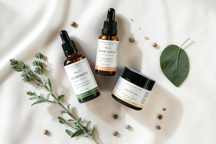 Apothecary & Me No.1 Illuminate Face Oil, No.2 Glow Face Scrub, and No.3 Calm Butter arranged on soft cream fabric with fresh herbs and a green leaf, a natural minimal-ingredient skincare range for supporting the skin barrier in psoriasis-prone skin