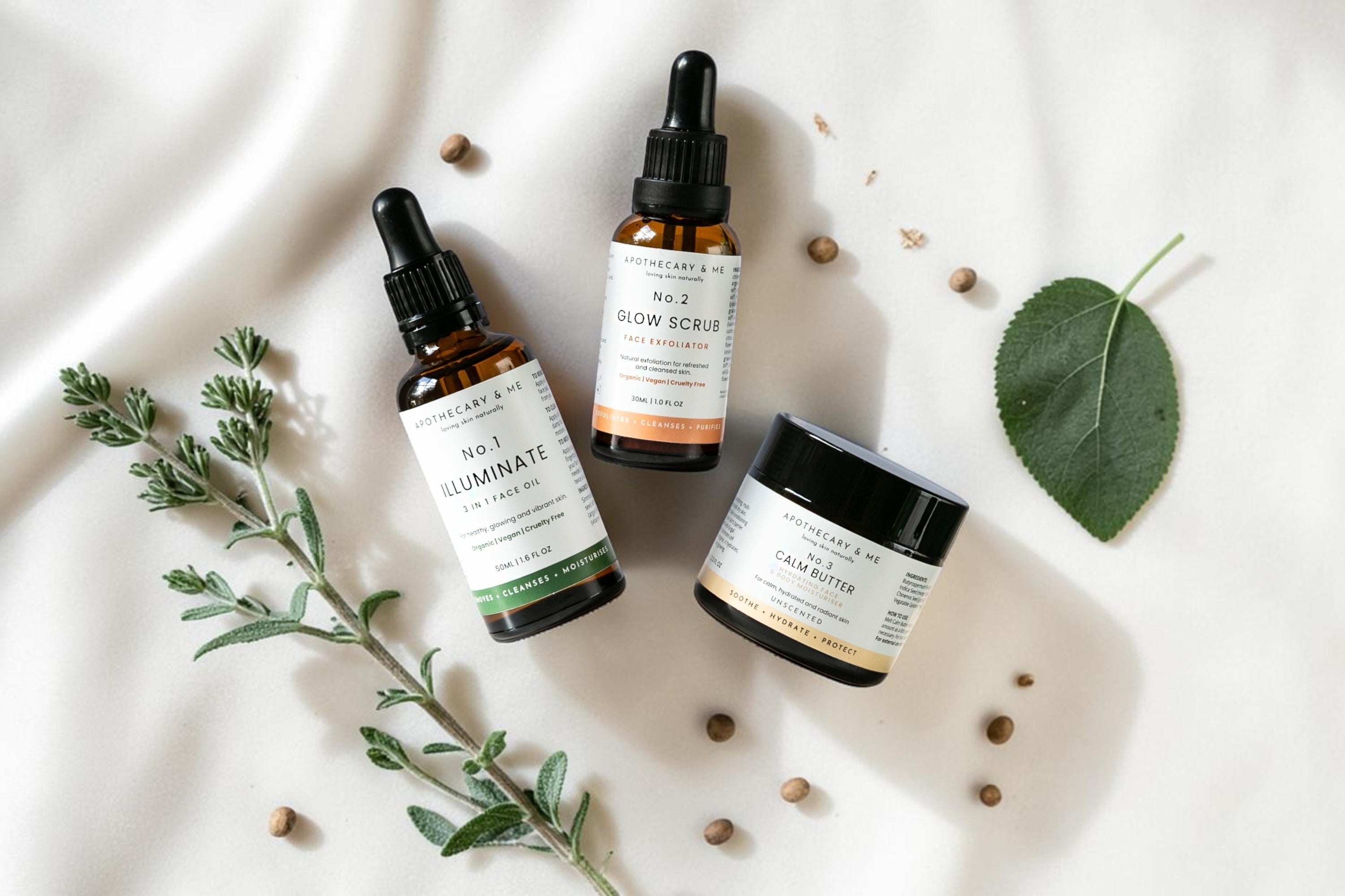 Apothecary & Me No.1 Illuminate Face Oil, No.2 Glow Face Scrub, and No.3 Calm Butter arranged on soft cream fabric with fresh herbs and a green leaf, a natural minimal-ingredient skincare range for supporting the skin barrier in psoriasis-prone skin