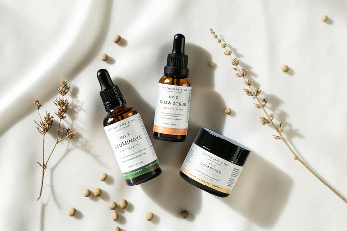  Apothecary & Me No.1 Illuminate Face Oil, No.2 Glow Face Scrub, and No.3 Calm Butter arranged on a soft cream fabric with dried botanicals, a minimal natural skincare routine for sensitive skin.