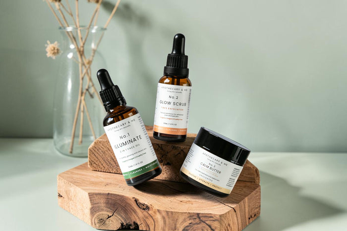  Apothecary & Me No.1 Illuminate Face Oil, No.2 Glow Face Scrub, and No.3 Calm Butter arranged on a raw wooden slice against a sage green background, a gentle minimal-ingredient natural skincare routine for topical steroid withdrawal.